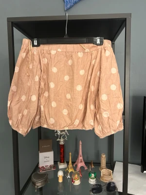 Beige prickig blus med puffärm och of shoulder  - Söt beige blus med vita prickar och stora puffärmar. Blusen har en avslappnad passform och resår vid ärmslut och nederkant. Perfekt för dig som gillar unika detaljer och ett lekfullt uttryck.