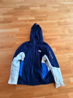Blå och vit vinterjacka från Helly Hansen - Vindjacka från Helly Hansen i marinblått med vita och blå detaljer på ärmarna. Jackan har huva, dragkedja framtill och logga på bröstet samt ärmen. Tillverkad i syntetmaterial och har långa ärmar.