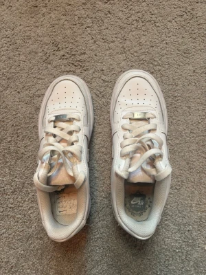 Vita Nike Air Force 1 sneakers - Klassiska vita Nike Air Force 1 sneakers i läder med perforerad tå, vita skosnören och den ikoniska swoosh-loggan på sidan. Låg modell med vadderad kant och robust sula som ger skön streetstil. Perfekta för dig som gillar tidlös och clean look.