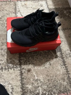 Svarta Nike Presto sneakers - Säljer ett par helsvarta Nike Presto sneakers med mesh-ovandel och elastisk passform. Skorna har en sportig design med rund tå, svarta snören och diskret Nike-logga. Perfekta för en streetwear-look och riktigt bekväma att ha på sig.