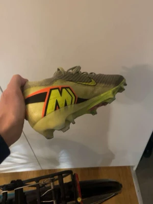 Nike Mercurial fotbollsskor gul - Säljer ett par Nike Mercurial fotbollsskor i gul färg med svarta och röda detaljer samt stort M på sidan. Skorna har snörning och är designade för bra grepp på planen. Lätta och smidiga med syntetmaterial för maximal bollkontroll.