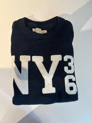 Mörkblå stickad tröja NY36 L.O.G.G. - Mörkblå stickad tröja från L.O.G.G. med stora vita bokstäver och siffror 'NY36' framtill. Rund halsringning och ribbstickade muddar. Perfekt för dig som gillar sportig och avslappnad stil. Köpt på HM. Se storlek på tröjan 