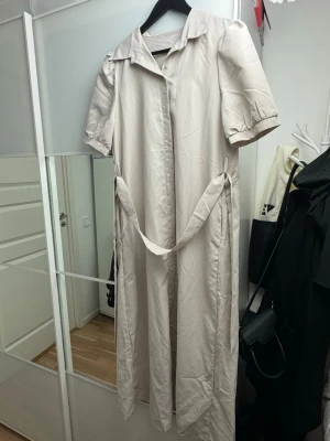 Beige långklänning med bälte - Stilren beige långklänning med korta puffärmar, knappar framtill och ett matchande bälte i midjan. Klänningen har klassisk krage och är tillverkad i ett mjukt bomullsliknande material. Perfekt för en avslappnad men ändå chic look.