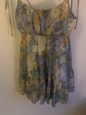 Blommig kortklänning med volanger - Söt kortklänning med tunna axelband och volangdetaljer. Klänningen har ett blommigt mönster i ljusa toner av blått, grönt, gult och vitt. Perfekt för varma dagar och har en luftig passform.