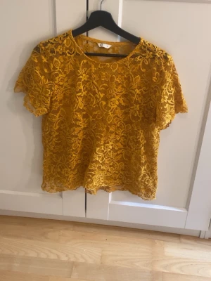 Gul spetsblus med blommönster - Superfin gul blus i spets med blommigt mönster och korta ärmar. Perfekt för dig som vill sticka ut med en färgglad och feminin topp. Blusen har rund halsringning och är lätt transparent tack vare spetsmaterialet.