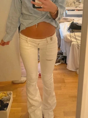 Lågmidjade jeans - Säljer ett par vita bootcut jeans med låg midja och snygga detaljer som fickor framtill och lätt slitning. Jeansen har en figurnära passform upptill och utsvängda ben. Perfekta för dig som gillar en trendig och avslappnad stil.
