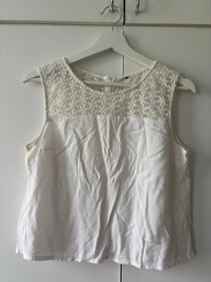 Vit blommig ärmlös blus med spets - Söt vit blus med ärmlös design och blommig spets upptill och på ryggen. Knäppning med knappar baktill och rund halsringning. Perfekt för en fräsch och somrig look.