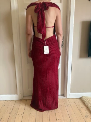 Vinröd långklänning med öppen rygg - Vinröd långklänning med v-ringning och öppen rygg. Klänningen har en snygg knytning i nacken och är figurnära med ett ribbat material. Perfekt för dig som vill sticka ut med en elegant och modern look. Aldrig använd och prislappen finns kvar.