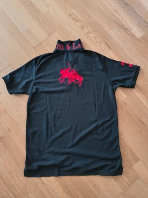 Svart piké från Ralph Lauren - Svart pikétröja från Ralph Lauren med röd broderad logga på bröstet och ryggen. Kragen har tydlig Ralph Lauren-text i rött och är försedd med knappar framtill. Kortärmad modell med siffran 3 broderad på ärmen. Klassisk och stilren design.
