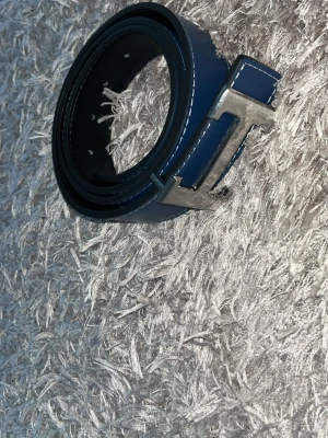 Blått skärp med silverfärgat spänne - Snyggt blått bälte med ett stort, silverfärgat spänne i form av ett H. Perfekt accessoar för att lyfta din outfit. Bältet har en stilren design och är gjort i ett slätt material.