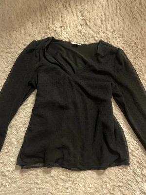 Svart omlottopp med prickigt mönster - Svart långärmad omlottopp med v-ringning och diskret prickigt mönster. Tunn och lätt genomskinlig, perfekt att styla lager på lager. Snygg passform och klassisk look.