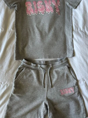 Grått set med tryck från Risky - Säljer ett grått tvådelat set från Risky med t-shirt och shorts. Båda delarna har ett rosa och vitt 'RISKY'-tryck framtill. Setet är gjort i mjukt bomullsmaterial och shortsen har dragsko i midjan samt fickor.