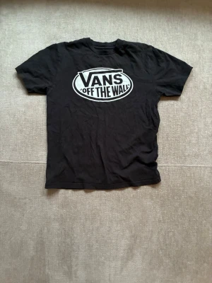 Svart Vans t-shirt med stor logga - Svart t-shirt från Vans med stor vit 'Vans Off The Wall'-logga på bröstet. Klassisk passform och rund halsringning, perfekt för en avslappnad streetstil. T-shirten är i mjuk bomull och har korta ärmar.