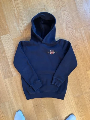 Mörkblå hoodie från GANT med brodyr - Snygg mörkblå hoodie från GANT med broderad logga och sköld på bröstet. Klassisk modell med huva och ribbade muddar vid ärmslut och nederkant. Perfekt för en avslappnad och stilren look.