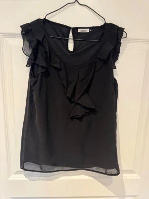 Svart volangblus från ONLY - Snygg svart ärmlös blus från ONLY med volangdetaljer längs axlar och bröst. Tillverkad i ett tunt, lätt material som ger ett luftigt intryck. Rund halsringning med liten knapp i nacken och dekorativ öppning bak.