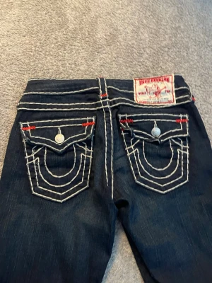 Mörkblå True Religion jeans med kontrastsömmar - Säljer ett par mörkblå jeans från True Religion med vita och röda kontrastsömmar. 