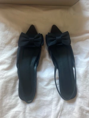 Svarta ballerinaskor med rosett - Snygga svarta ballerinaskor med spetsig tå och stor dekorativ rosett framtill. Skorna har öppen häl med smal rem och är tillverkade i ett slätt material som ger en elegant look. Perfekta för dig som gillar stilrena detaljer och trendig design.