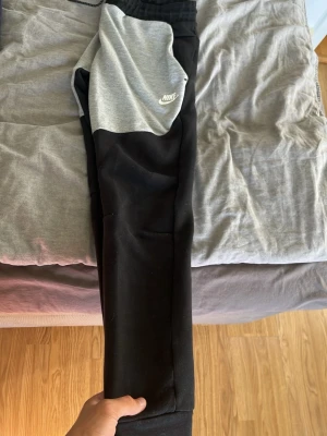 Nike svart-grå mjukisbyxor - Nike mjukisbyxor i svart med grå paneler upptill och vit Nike-logga på vänster sida. Byxorna har resår i midjan med dragsko och smal passform. Perfekta för chill eller träning, tillverkade i mjukt material för extra komfort.