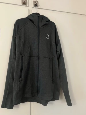 Mörkgrå hoodie från Haglöfs - Mörkgrå hoodie från Haglöfs med dragkedja hela vägen, två fickor med dragkedja och diskret logga på bröstet. Tillverkad i mjukt och stretchigt material, perfekt för en aktiv livsstil. Snygg och enkel design med huva.