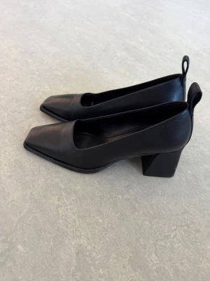 Svarta pumps med fyrkantig tå - Stilrena svarta pumps från Vagabond i skinn med fyrkantig tå och bred klack. Skorna har en modern design med dragflik baktill och passar perfekt till dig som gillar minimalistisk stil.