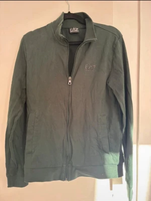 Grön zip-hoodie från EA7 Emporio Armani - Snygg grön hoodie med dragkedja från EA7 Emporio Armani. Tröjan har ståkrage, två fickor framtill och diskret logga på bröstet. Tillverkad i mjukt bomullsmaterial, perfekt för en avslappnad och trendig stil.