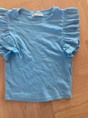 Blå ribbad topp med volangärm Zara - Supersöt ljusblå ribbad topp från Zara med volangärmar. T-shirten har rund halsringning och är tillverkad i ett mjukt, stretchigt material som sitter snyggt på kroppen. Perfekt för dig som vill ha en trendig och bekväm look.