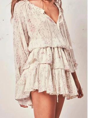 Blommig volangklänning från LoveShackFancy - Romantisk kort klänning i ljusbeige med rosa och lila blommönster från LoveShackFancy. Klänningen har långa ärmar, volangkjol i flera lager, v-ringning med knytband och resår i midjan. Tillverkad i ett tunt, luftigt tyg som ger en bohemisk känsla. Pasar bra på s storlek 