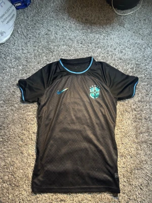 Svart Brasilien fotbollströja Nike - Snygg svart fotbollströja från Nike med Brasilien CBF-logga och turkosa detaljer vid krage och ärmslut. Tröjan har korta ärmar och är tillverkad i ett lätt och ventilerande material, perfekt för träning eller match.