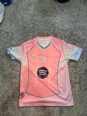 Barça fotbollströja Lamine Yamal #10 - Säljer en FC Barcelona fotbollströja från Nike i rosa och ljusblått med Lamine Yamal och nummer 10 på ryggen. Tröjan har korta ärmar, V-ringning och klubbmärke samt sponsorlogga på bröstet. Perfekt för fotbollsälskare som vill sticka ut på planen.