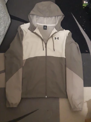 Under Armour vindjacka med huva - Stilren vindjacka från Under Armour i beige och ljusbrun färgblock-design. Jackan har huva med dragsko, hel dragkedja och elastiska muddar vid ärmslut. Perfekt för blåsiga dagar och sportiga outfits.