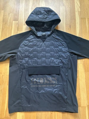 BOSS J_Liu Hybridjacka - (Oanvänd) Passar M/L - Svart vindjacka från BOSS med quiltad front, stor magficka och tydlig logga. Jackan har huva, raglanärmar och elastiska muddar. Materialet är en mix av slätt och quiltat tyg som ger en sportig och modern känsla. Helt oanvänd men utan tags. Nypris: Ca 3700 kr. Jackan passar för dem som bär M/L. Hör av dig för mer info. 