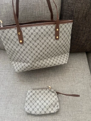 Beige handväska  - Snygg beige handväska med klassiskt monogrammönster och bruna läderdetaljer. Väskan har två axelband i brunt läder med guldfärgade nitar och matchande liten necessär med dragkedja. Perfekt för dig som gillar stilrena accessoarer. Pris går såklart att diskutera hör av er !