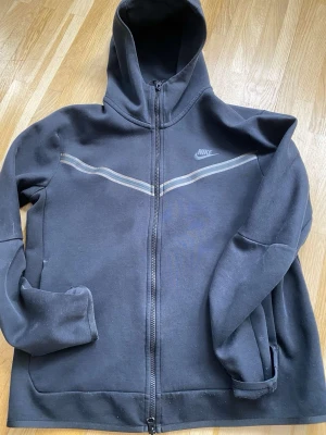 Svart Nike hoodie med dragkedja - Säljer en svart nike tech fleece väldigt skönt och snygg. Lite sol blekt på ränderna på brösten och har ett litet hål nere på ena armen 