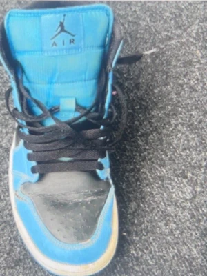 Blåa Air Jordan 1 High sneakers - Snygga Air Jordan 1 High sneakers i blått och svart med klassisk Nike Swoosh på sidan och Air Jordan-logga vid ankeln. Skorna har svarta skosnören och vit sula med blå detaljer. Ovandelen är i läder och textil för en sportig look.