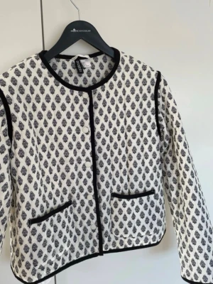 Mönstrad cardiganjacka från H&M - Snygg cardiganjacka från H&M i vitt med svart bladliknande mönster. Jackan har rund halsringning, svarta kantband och två framfickor. Långärmad och i mjukt, vävt material som ger en stilren look.
