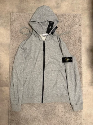 Grå hoodie från Stone Island - Snygg grå hoodie från Stone Island med dragkedja framtill och klassisk logopatch på vänster ärm. Tillverkad i mjukt bomullsmaterial och har huva med dragsko samt ribbade muddar. Perfekt för en avslappnad och trendig stil.