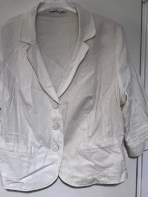 Vit kavaj med tre knappar - Stilren vit kavaj med tre knappar framtill och två fickor. Kavajen har klassisk krage och 3/4-ärmar. Perfekt för en fräsch och clean look. Materialet ser ut att vara bomull eller linneblandning.