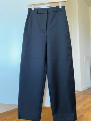 Svarta vida kostymbyxor - Stilrena svarta kostymbyxor med hög midja och raka, vida ben. Byxorna har hällor för skärp och klassisk stängning fram. Perfekta för en dressad look och enkla att matcha med olika toppar.