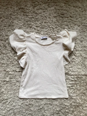 Vit topp med volangärmar från Zara - Supersöt vit topp från Zara med volangärmar och rund halsringning. Toppen är ribbad och har en lätt croppad passform. Perfekt att styla med jeans eller kjol för en trendig look. Aldrig använd och är som ny!