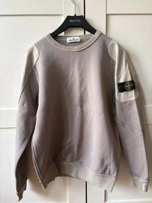 Grön/beige sweatshirt från Stone Island - Säljer en grön/beige sweatshirt från Stone Island med klassisk rund hals och ribbade muddar. Tröjan har ikonisk svart patch med gult och grönt Stone Island-märke på vänster ärm. Tillverkad i mjuk bomull. 