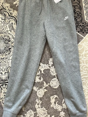 Oanvänd mjukisbyxa från Nike - Snygga grå mjukisbyxor från Nike med dragsko i midjan och ribbade muddar vid bensluten. Klassisk Nike-logga broderad på vänster sida. Tillverkade i mjukt och bekvämt material som passar perfekt för chill eller träning.
