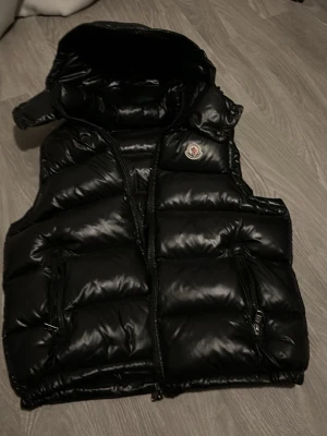 Svart dunväst från Moncler - Svart dunväst från Moncler med glansig finish och avtagbar huva. Västen har två stora fickor med dragkedja och klassisk Moncler-logga på bröstet. Perfekt för lager-på-lager och streetwear-vibe.