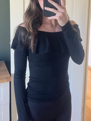 Svart offshoulder topp med volang - Trendig svart offshoulder topp med långa ärmar och volangdetalj över bysten. Sitter tight och har en stilren, enkel design som passar perfekt till jeans eller kjol. Perfekt för dig som gillar en stilren men ändå lite extra look.