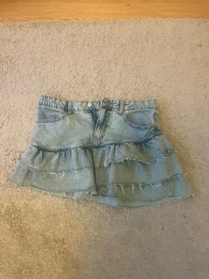 Ljusblå jeanskjol med volanger - Supersnygg ljusblå jeanskjol från perfect jeans i storlek 38. Kjolen har två volanglager och råa, fransiga kanter för en trendig look. Klassisk femficksmodell med knapp och dragkedja framtill. Perfekt till sommaren!
