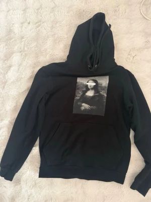 Svart hoodie med Mona Lisa-tryck - Svart hoodie med stor ficka framtill och tryck av Mona Lisa på bröstet. Klassisk huva med dragsko och ribbade muddar. Liten etikett på ryggen upptill. Perfekt för dig som gillar streetwear och konstinspirerade plagg.