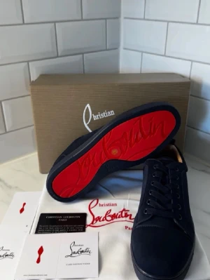Mörkblå sneakers från Christian Louboutin - Snygga mörkblå sneakers från Christian Louboutin med klassisk röd sula och broderad logga på hälen. Skorna har en stilren design i mocka och snörning framtill. Perfekta för dig som vill sticka ut med exklusiva detaljer.