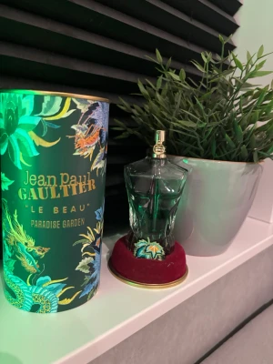 Jean Paul Gaultier Le Beau Paradise Garden - Unik parfymflaska formad som en manlig torso i grönt glas med gulddetaljer. Kommer med en dekorativ grön och guldig förpackning med tropiskt mönster och texten 'Le Beau Paradise Garden'. Perfekt för dig som gillar exklusiva dofter och snygg design.