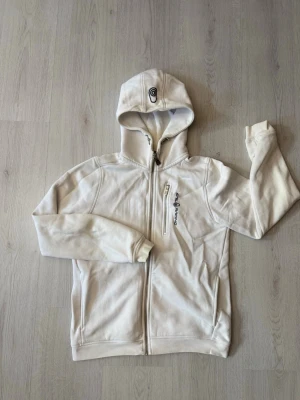 Vit hoodie från Sail Racing  - Säljer en stilren vit hoodie från Sail Racing med dragkedja framtill och broderad logga på huvan. Hoodien har fickor med dragkedja och är tillverkad i mjukt bomullsmaterial. Perfekt för en avslappnad look och enkel att matcha. Har tyvärr en fläck på ärmen.