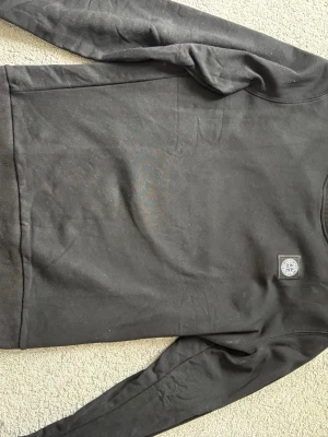 Svart sweatshirt från Stone Island - Svart sweatshirt från Stone Island med klassisk rund hals och ribbade muddar. Tröjan har den ikoniska Stone Island-loggan broderad på bröstet. Perfekt för en stilren och avslappnad look.