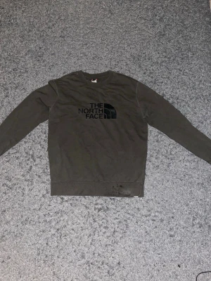 Olivgrön sweatshirt från The North Face - Olivgrön sweatshirt med svart The North Face-logga på bröstet. Klassisk rund halsringning och långa ärmar. Tröjan har ribbade muddar vid ärmslut och nederkant. Perfekt för en avslappnad och stilren look.